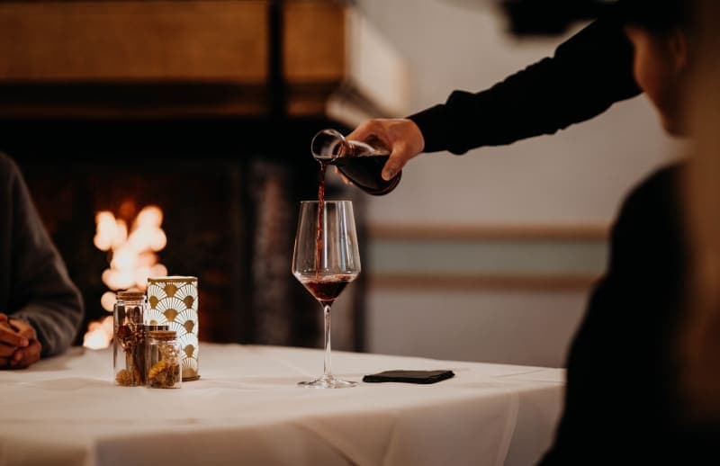 Kellner schenkt Rotwein in ein Glas am elegant gedeckten Tisch vor einem brennenden Kamin